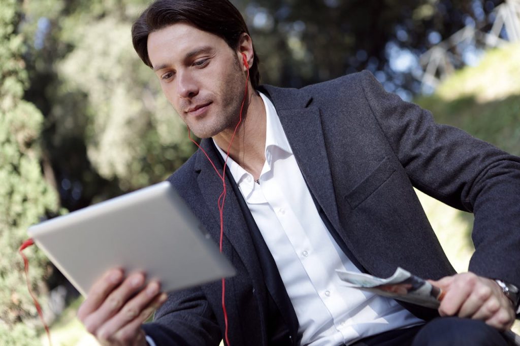 Businessman in suit using a tablet with headphones in a park setting, exemplifying mobile technology use.